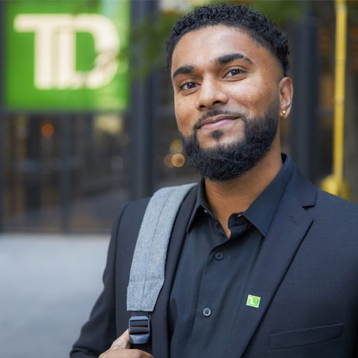 Un étudiant souriant tient un ordinateur portable et un téléphone à l’extérieur d’un campus collégial.