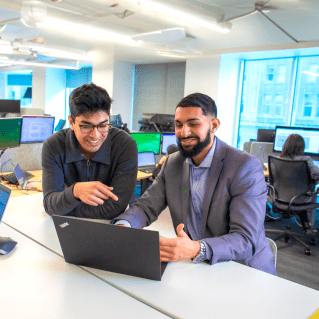 Two colleagues smile while looking at a laptop screen together.
