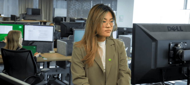 A new colleague wearing a blazer and green TD pin works on a computer in a busy office.