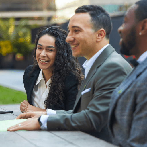 Trois collègues assis à une table à l’extérieur sourient.